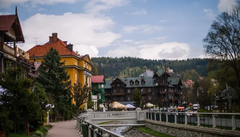 Urokliwa promenada w Krynicy-Zdroju, z widokiem na zabytkowe wille i górskie zbocza. W oddali widać budynek z napisem "Polonus", przypominający o podróży z Warszawy Zachodniej.