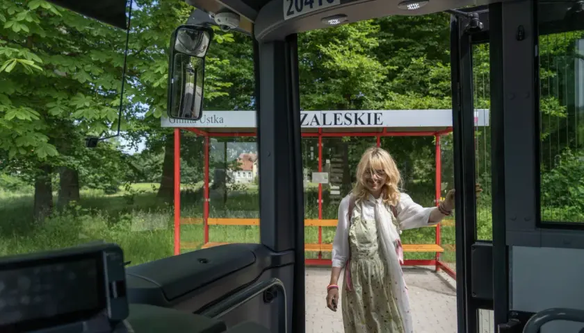 Młoda kobieta wsiada do autobusu na przystanku w gminie Ustka. W tle zielony krajobraz i budynek.