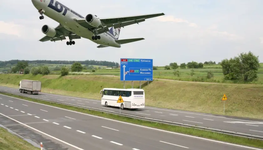 Samolot LOT leci nisko nad autostradą A4 w kierunku Katowic. Obok jedzie autobus, a w oddali widać drogę do Krakowa i Warszawy.