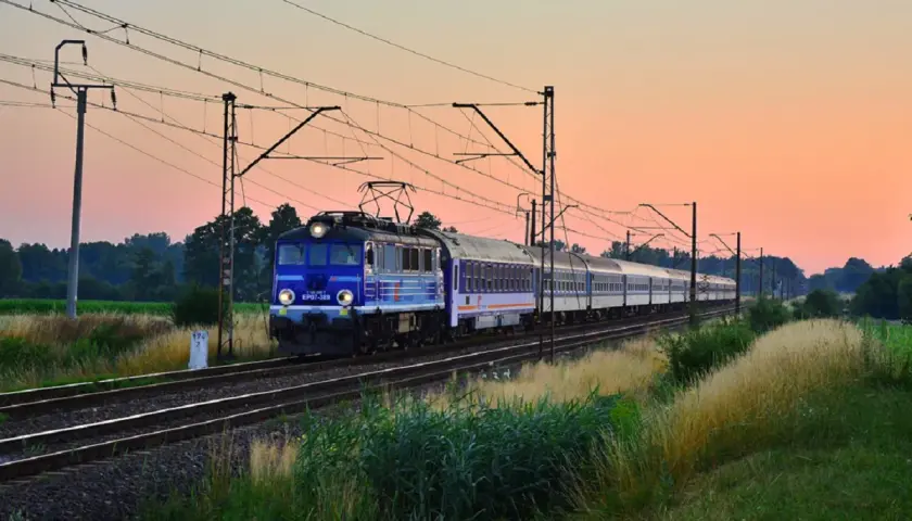 Niebieski pociąg PKP EP07-389 z wagonami mija pole o zachodzie słońca, jadąc w stronę Kołobrzegu.