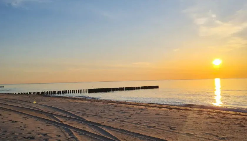 Zachód słońca nad morzem, piaszczysta plaża z widocznymi śladami opon, falochrony. Idealne miejsce na odpoczynek po podróży, nawet jeśli wicie dojazd PKP.
