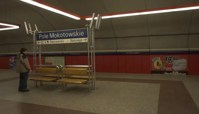 Na stacji metra Pole Mokotowskie stoi mężczyzna obok ławki. W tle niebieska tablica z nazwami ulic i czerwone panele.