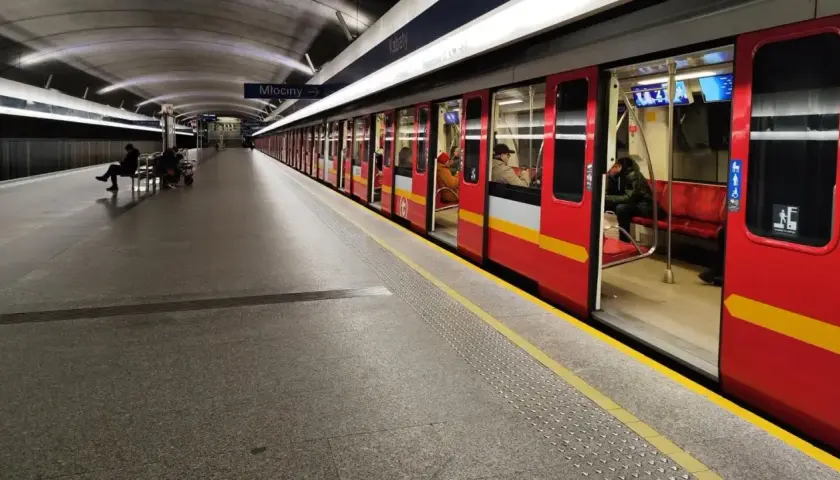 Czerwony pociąg metra M1 z otwartymi drzwiami na stacji. Pasażerowie czekają na peronie.