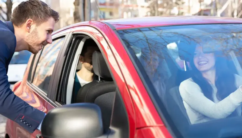 Mężczyzna w garniturze pochyla się do czerwonego samochodu, by porozmawiać z uśmiechniętą kobietą za kierownicą. To świetny przykład carpoolingu!