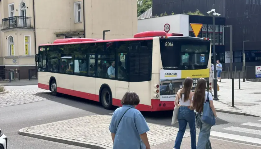 Autobus linii 148 w Gdańsku, z numerem bocznym 8016, odjeżdża z przystanku. Na pierwszym planie dwie dziewczyny idące przez pasy.