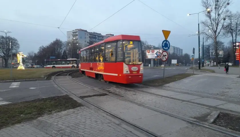 Czerwony ztm 26 tramwaj przejeżdża przez skrzyżowanie. W tle widać autobus i budynki.