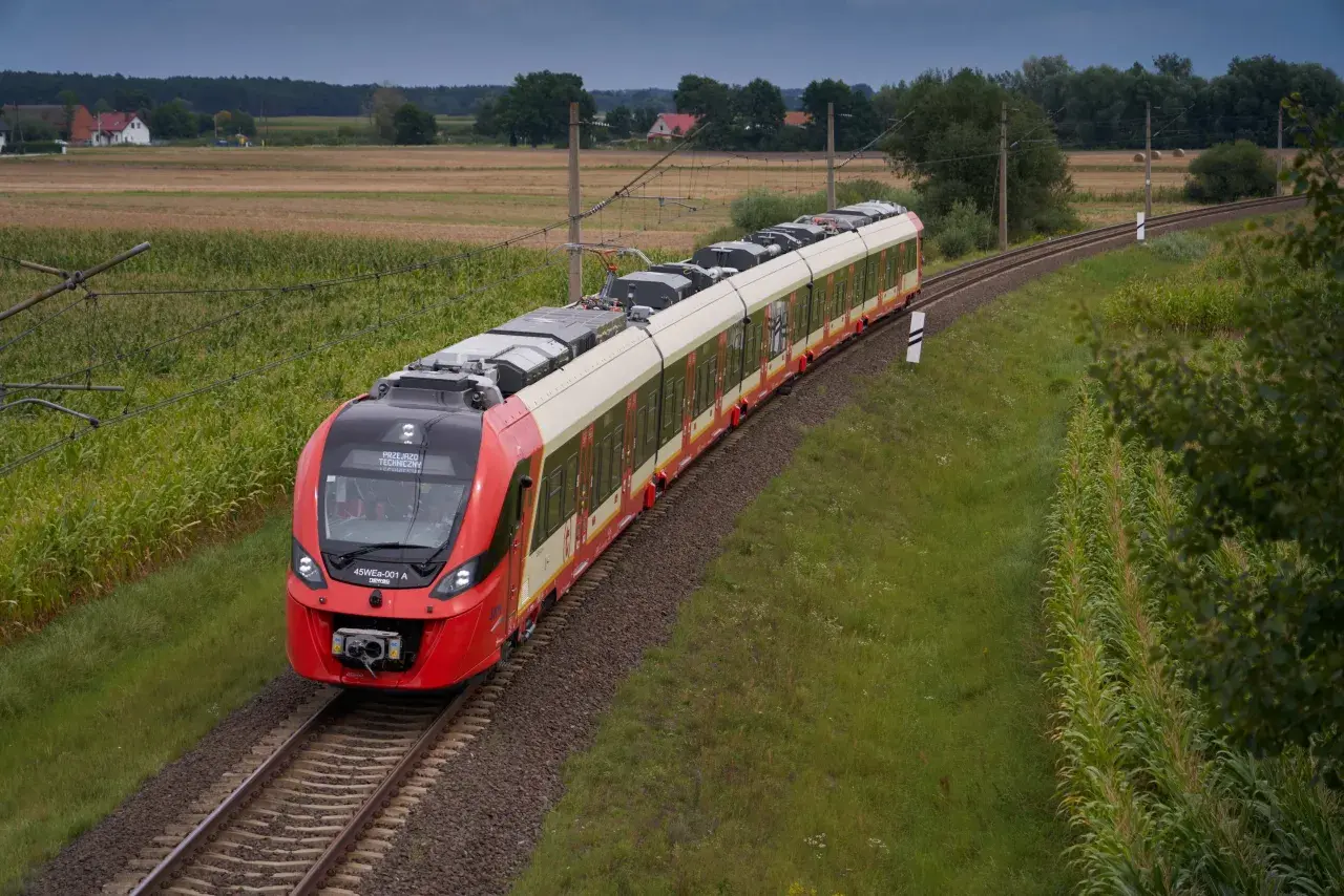 Czerwony pociąg SKM jedzie po torach między polami kukurydzy i zboża, w tle domy i las. Trasa Warszawa-Legionowo.