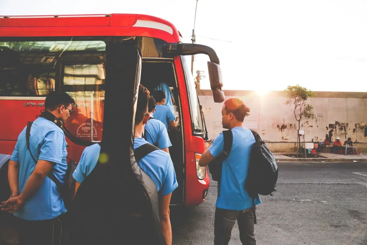 Grupa młodych ludzi z gitarą wsiada do czerwonego autobusu GTV. Słońce zachodzi, tworząc ciepłą atmosferę.