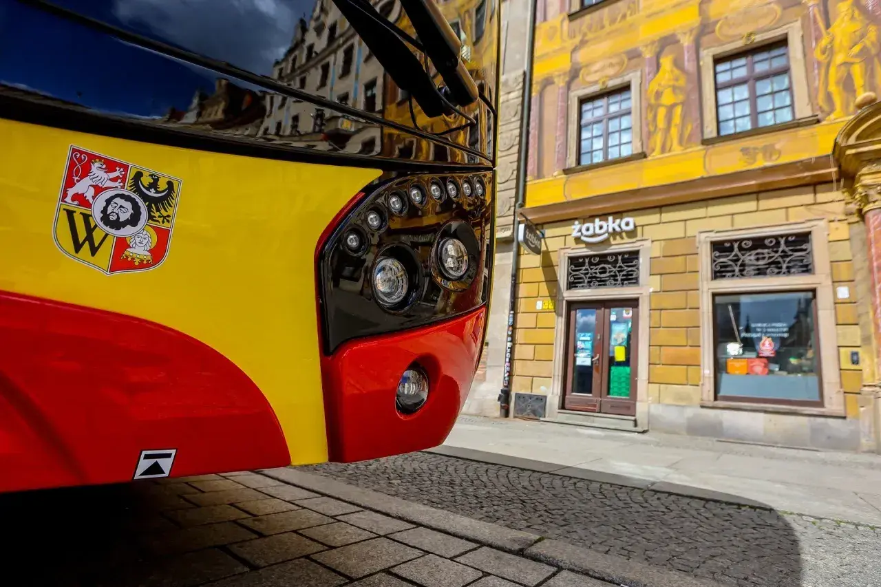 Autobus kraków komunikacja miejska z herbem miasta. Na żółto-czerwonym pojeździe widoczny herb Krakowa. W tle kamienica z malowidłami i sklep Żabka.
