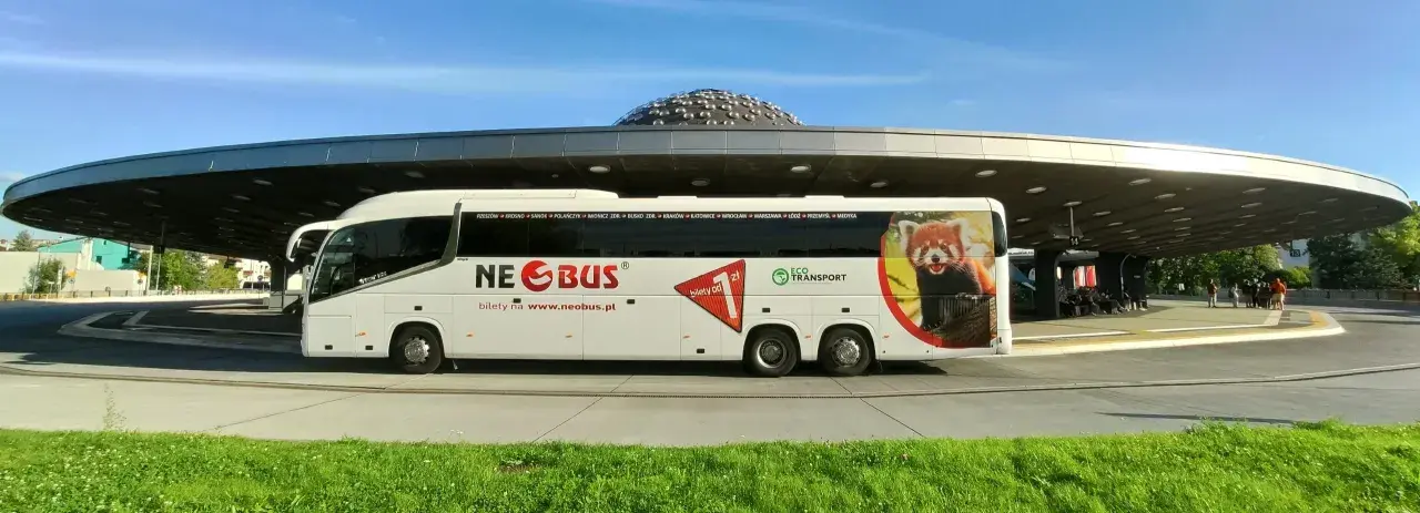 Biały autokar Neobus z pandą małą na boku stoi pod nowoczesnym zadaszeniem. Sprawdź neobus ceny biletów na www.neobus.pl.