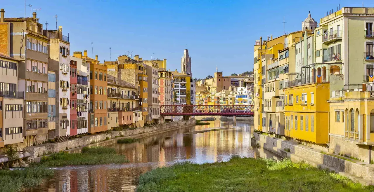 Kolorowe kamienice nad rzeką w Gironie, z mostem łączącym z Barceloną w tle.