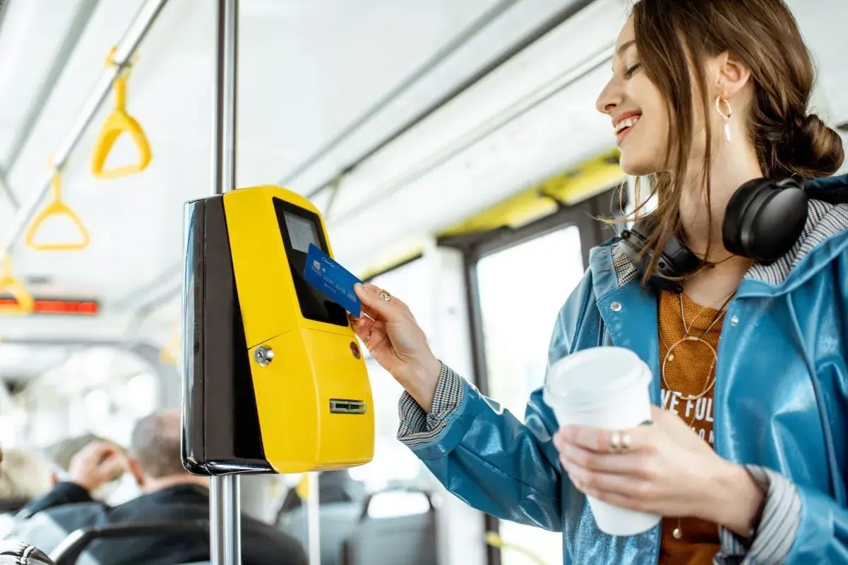 Uśmiechnięta kobieta z kawą w ręku zbliża kartę do żółtego kasownika, aby aktywować swój bilet okresowy w autobusie.