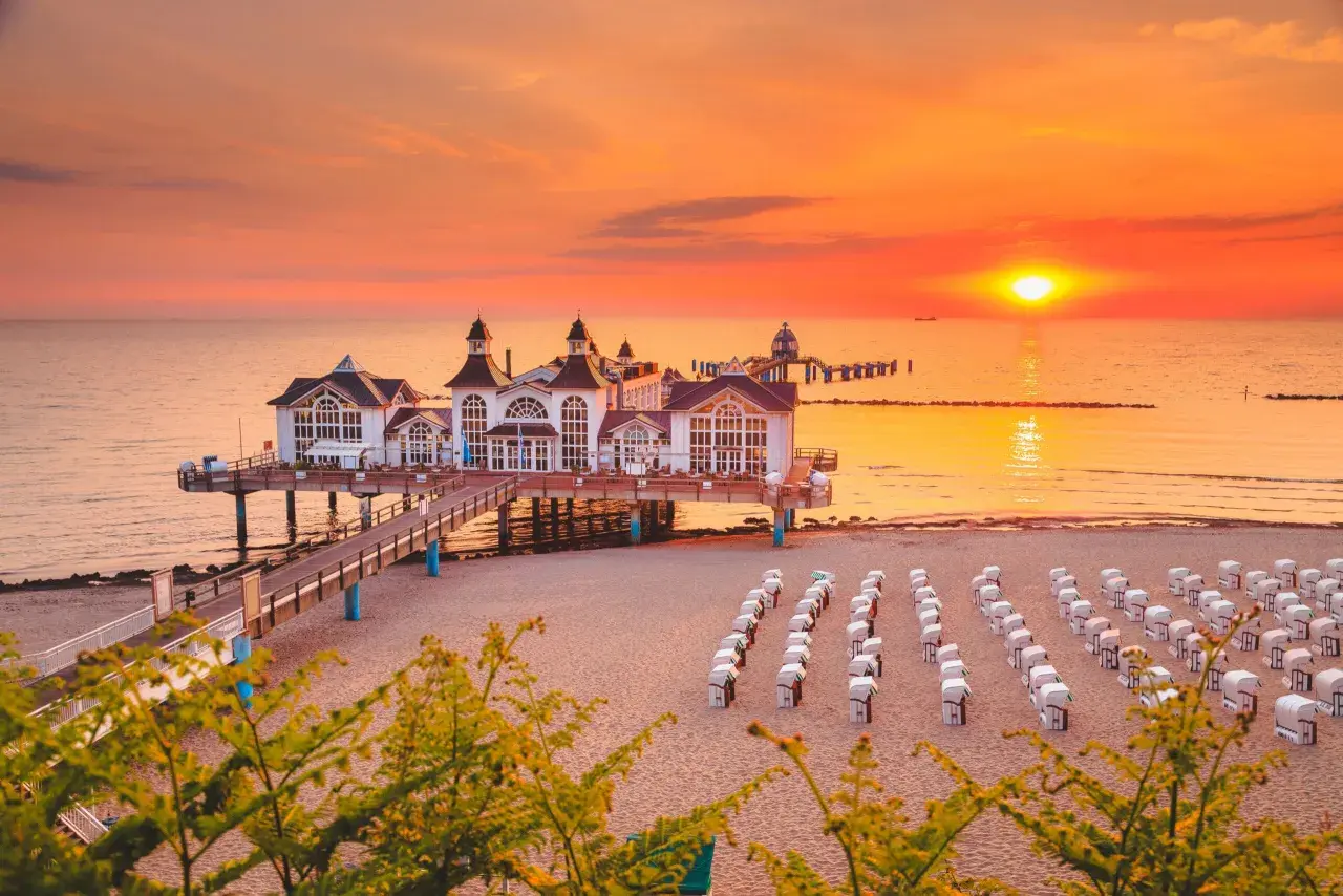 Zachód słońca nad Rugią, idealny cel podróży z Polski. Molo z białymi budynkami, plaża z rzędami koszy plażowych.