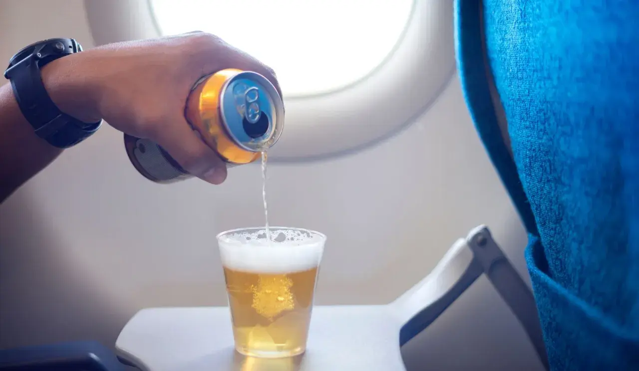 Ręka z puszką piwa nalewa napój do szklanki na stoliku w samolocie. Czy można przewozić alkohol samolotem?