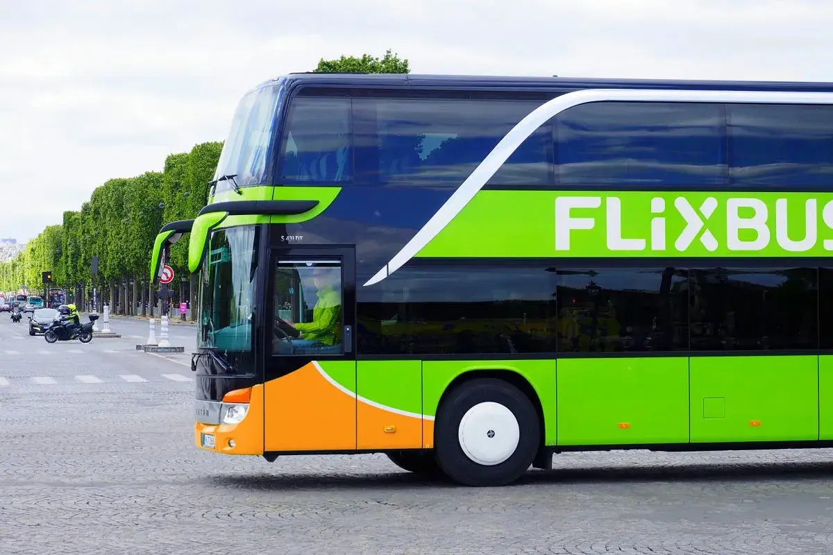 Zielony autokar Flixbus z pomarańczowym pasem na dole, jadący ulicą. Kierowca w żółtej kurtce. Czy Flixbus robi postoje? Tak, ten autokar jest w ruchu.