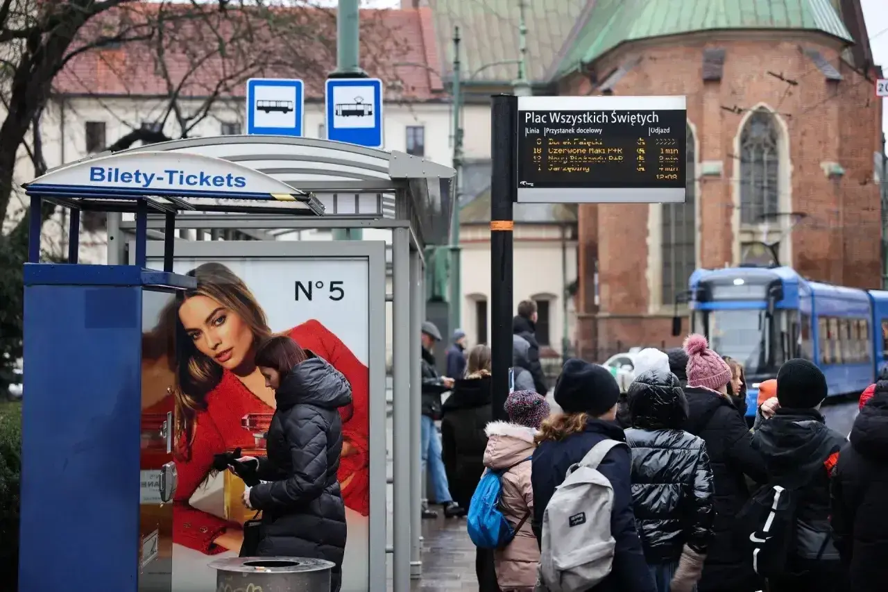 Kraków, Plac Wszystkich Świętych. Ludzie czekają na tramwaj, obok automat z biletami. W tle zabytkowa architektura i niebieski tramwaj.