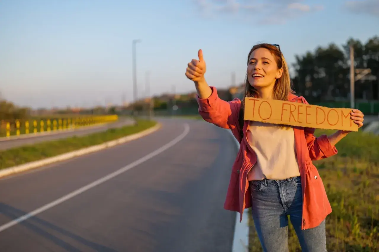 Młoda kobieta z uśmiechem na twarzy łapie autostop. Trzyma tabliczkę "TO FREEDOM" i kciuk w górę, gotowa na przygodę.