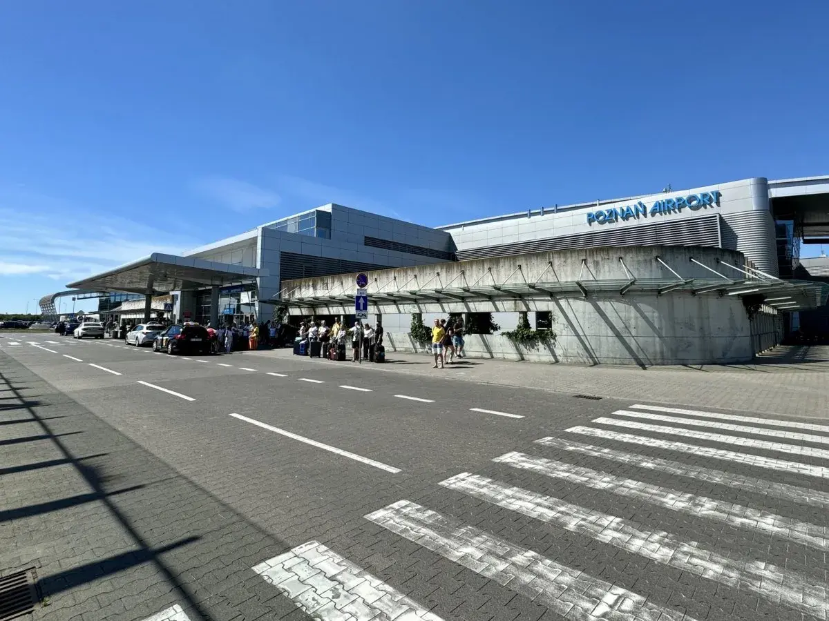 Lotnisko Poznań Airport, z ludźmi i samochodami przed budynkiem. Tu znajdziesz informacje, jak dojechać z dworca PKP na lotnisko w Poznaniu.