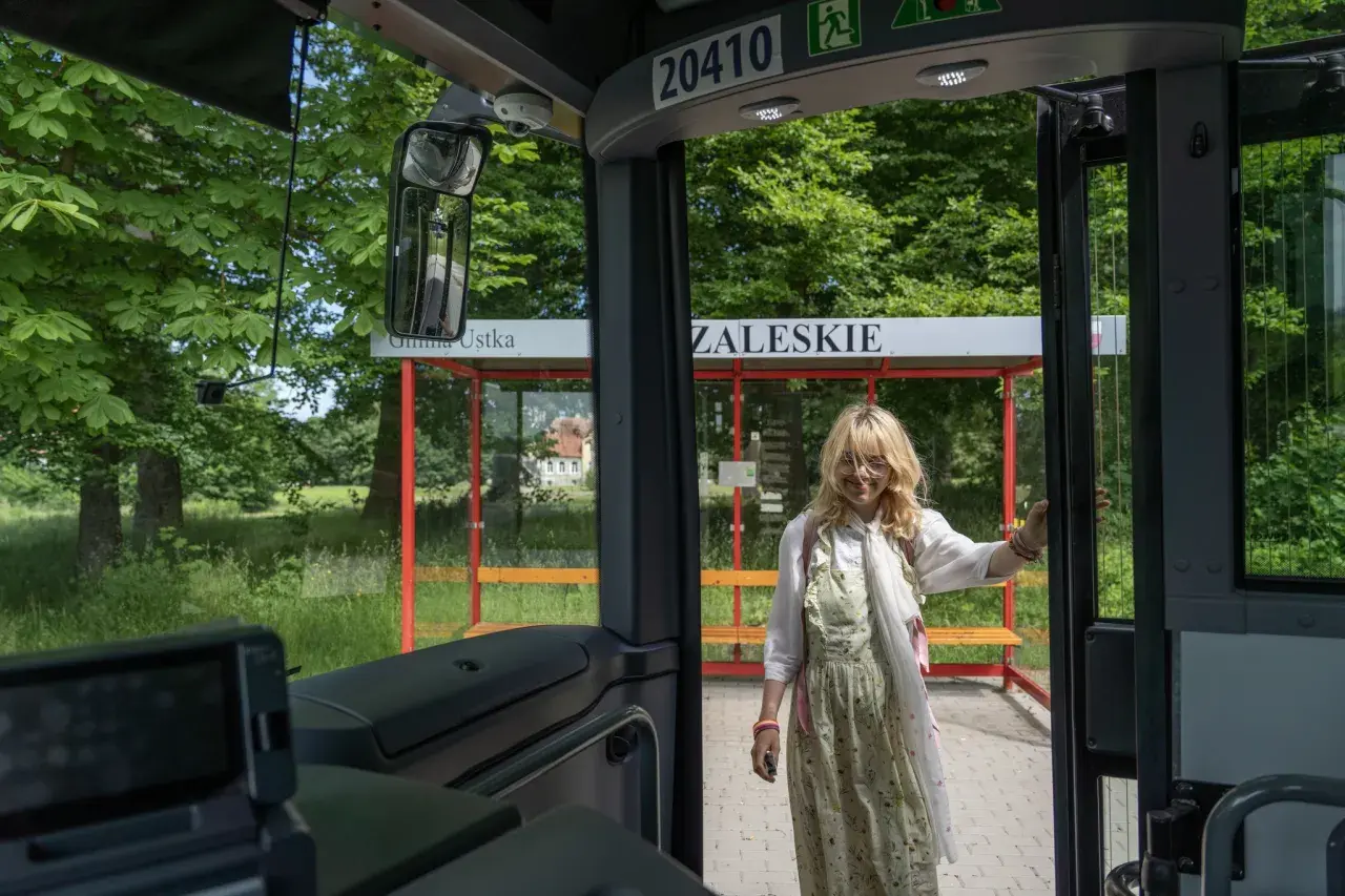 Młoda kobieta wsiada do autobusu na przystanku w gminie Ustka. W tle zielony krajobraz i budynek.