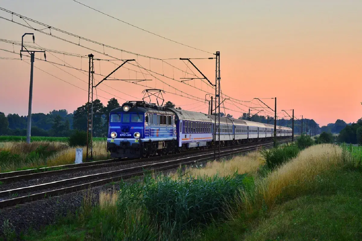 Niebieski pociąg PKP EP07-389 z wagonami mija pole o zachodzie słońca, jadąc w stronę Kołobrzegu.