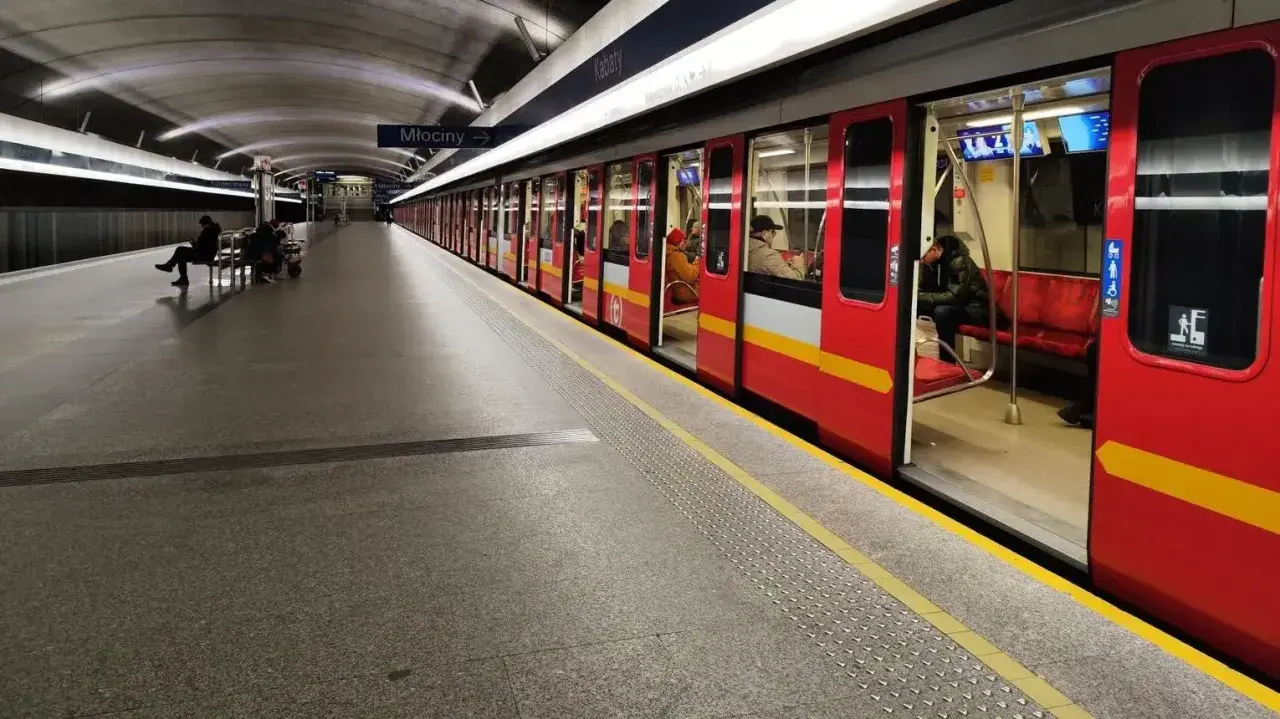 Czerwony pociąg metra M1 z otwartymi drzwiami na stacji. Pasażerowie czekają na peronie.