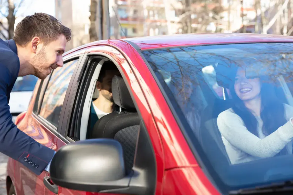 Mężczyzna w garniturze pochyla się do czerwonego samochodu, by porozmawiać z uśmiechniętą kobietą za kierownicą. To świetny przykład carpoolingu!
