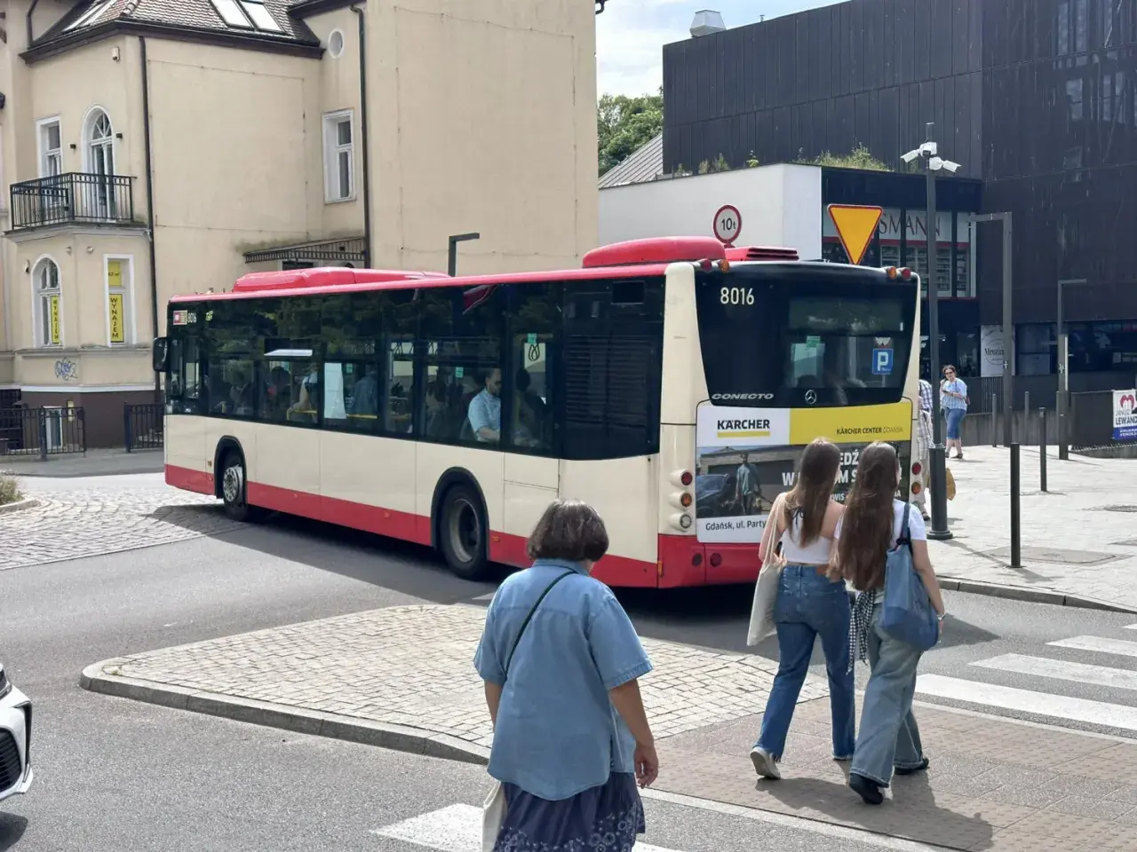 Autobus linii 148 w Gdańsku, z numerem bocznym 8016, odjeżdża z przystanku. Na pierwszym planie dwie dziewczyny idące przez pasy.