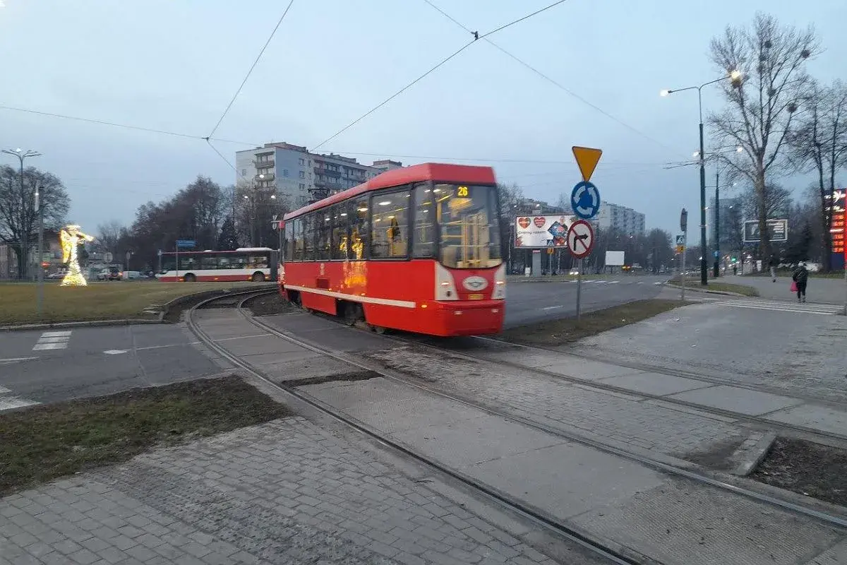 Czerwony ztm 26 tramwaj przejeżdża przez skrzyżowanie. W tle widać autobus i budynki.