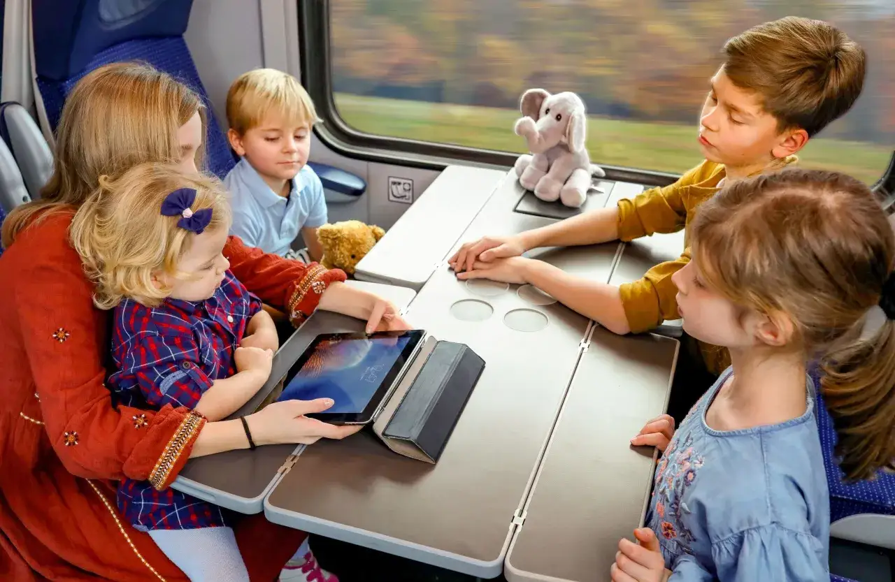 Rodzina z dziećmi podróżuje pociągiem PKP. Dzieci oglądają bajki na tablecie, a mama trzyma malucha na kolanach.