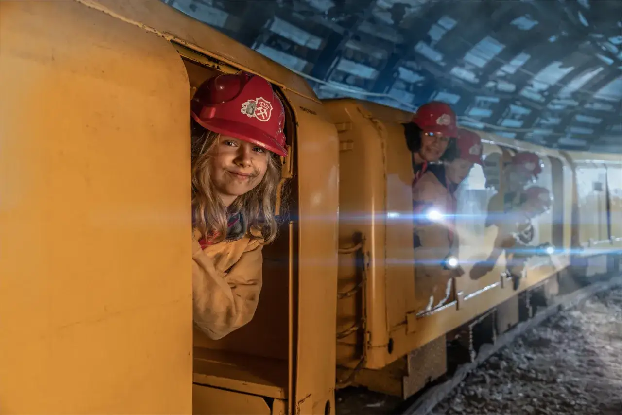 Uśmiechnięta dziewczynka w kasku w żółtym wagonie, gotowa na jednodniową wycieczkę pociągiem. Inni pasażerowie z latarkami w kolejnych wagonach.