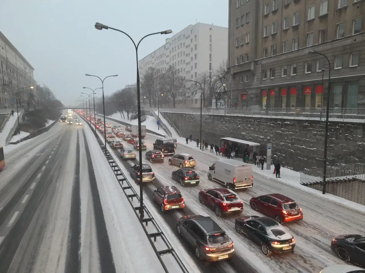Zimowy korek na Bemowie, samochody stoją w śniegu. W tle widać budynki i przystanek autobusowy.