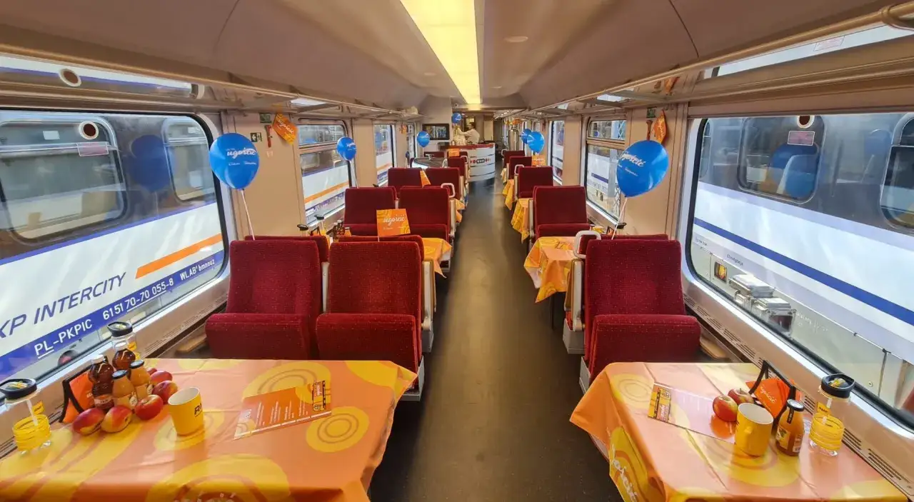 Wnętrze wagonu Pendolino przygotowane na przyjęcie gości. Na stolikach o pomarańczowych obrusach stoją napoje i owoce. Błękitne balony dodają uroku.