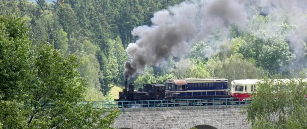 Parowóz z wagonami przemierza kamienny most, dymiąc obficie. To malownicza podróż pociągiem Szklarska Poręba Harrachov przez zielone Karkonosze.