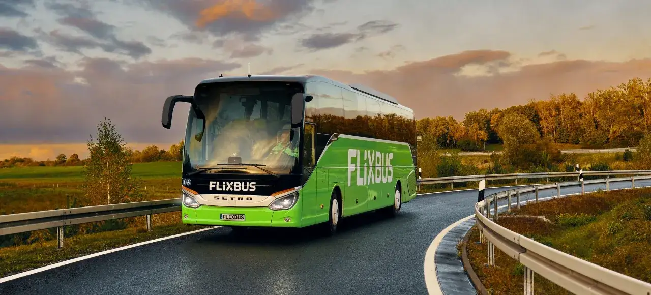 Zielony autobus Flixbus jedzie po krętej drodze wśród jesiennych drzew. Szukaj flixbus promocje na podróże!