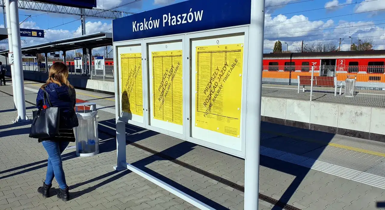 Kobieta czeka na pociąg na stacji Kraków Płaszów, do której jeździ metro. Na tablicy widnieje rozkład jazdy.
