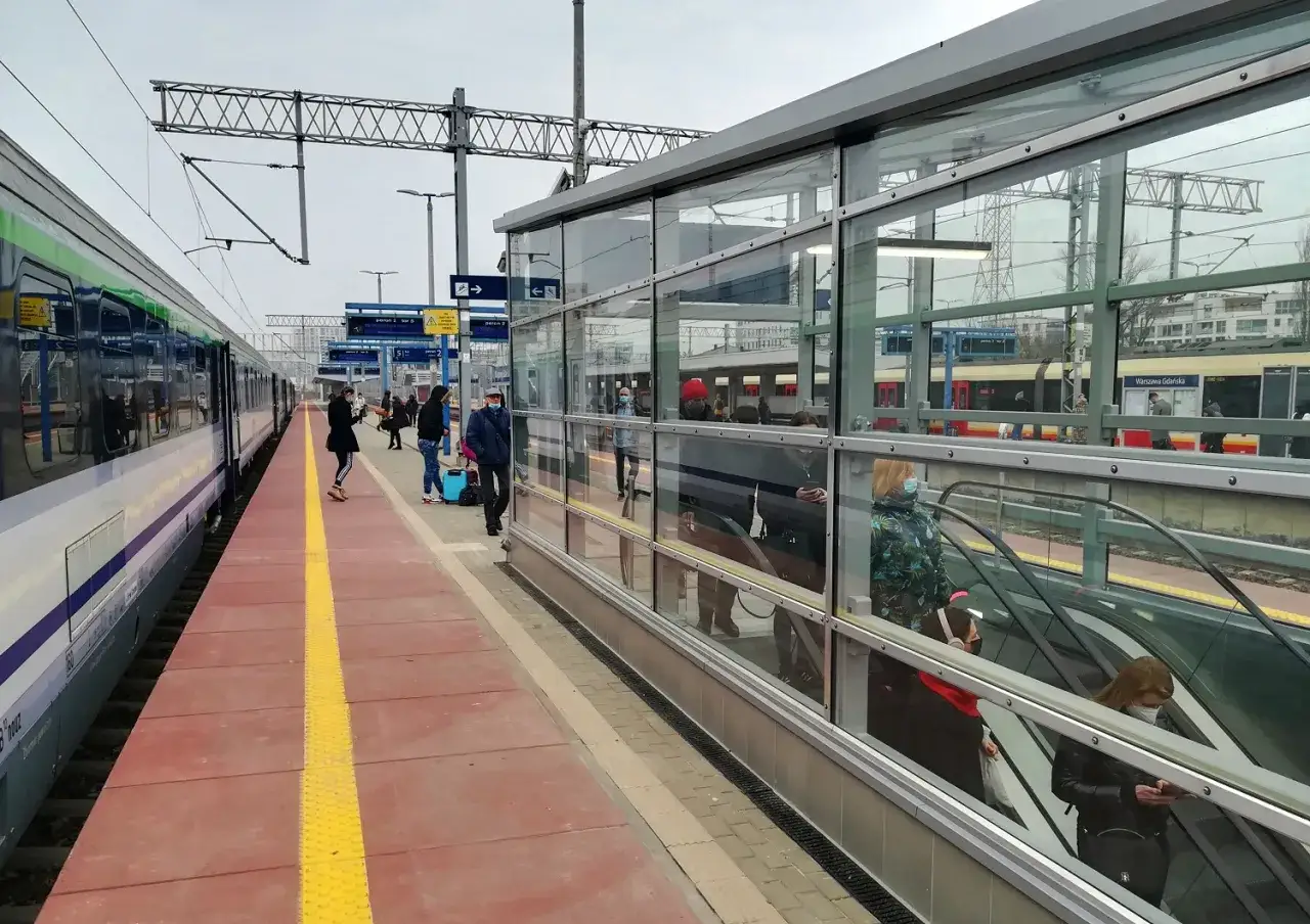 Pociąg Warszawa - Gdańsk czeka na pasażerów. Na peronie widać ludzi z bagażami, a w tle nowoczesna hala z ruchomymi schodami.