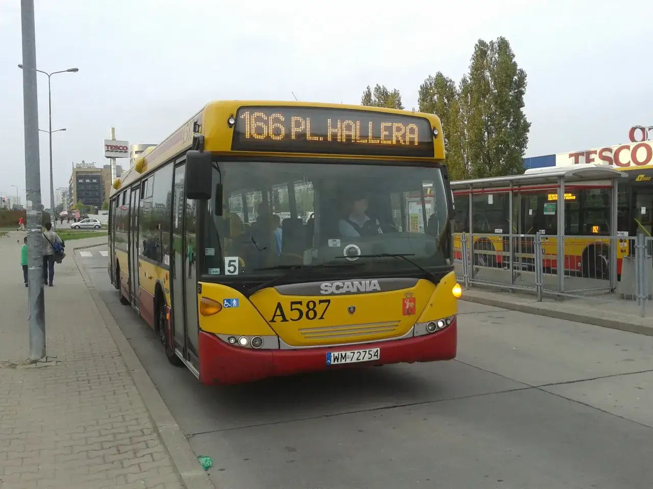 Żółty autobus linii 166 w Warszawie, z numerem bocznym A587, zbliża się do przystanku obok Tesco.