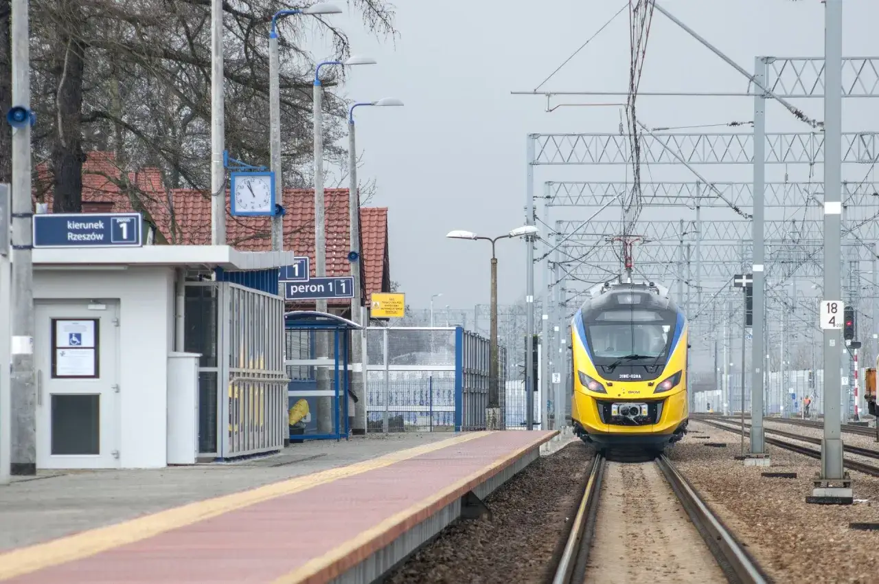 Żółty pociąg zbliża się do peronu 1. Na peronie widać wiatę i tablicę z kierunkiem Rzeszów.