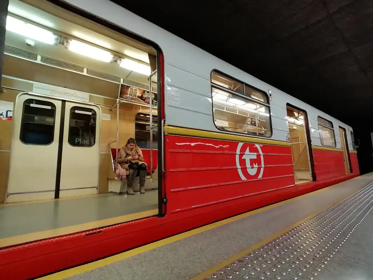 Wnętrze czerwono-białego metra, gdzie jedna pasażerka czeka na peronie. To świąteczne metro jest gotowe na podróżnych.