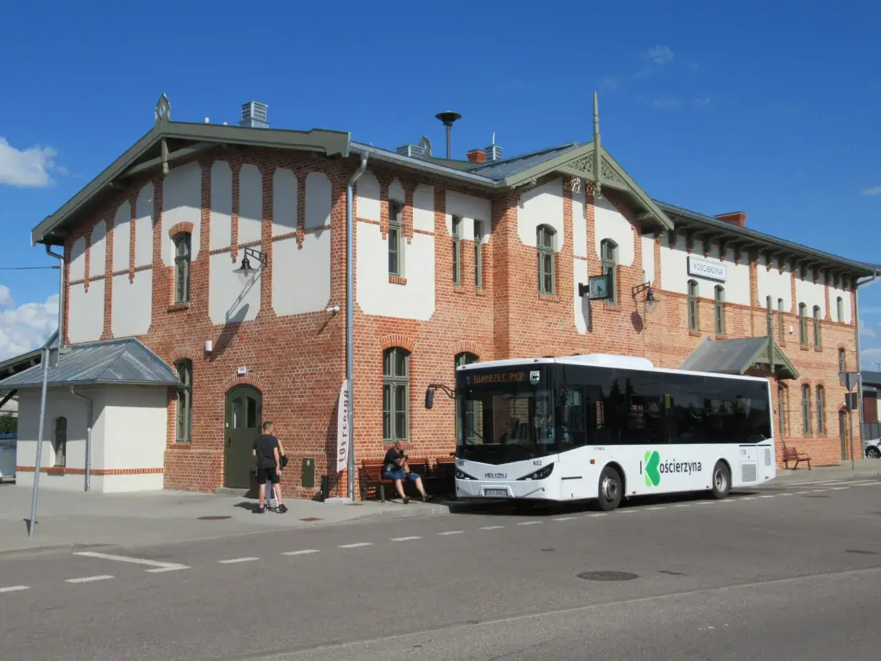 Autobusowa komunikacja zastępcza w Kościerzynie. Biały autobus marki Isuzu stoi przed ceglanym budynkiem dworca.