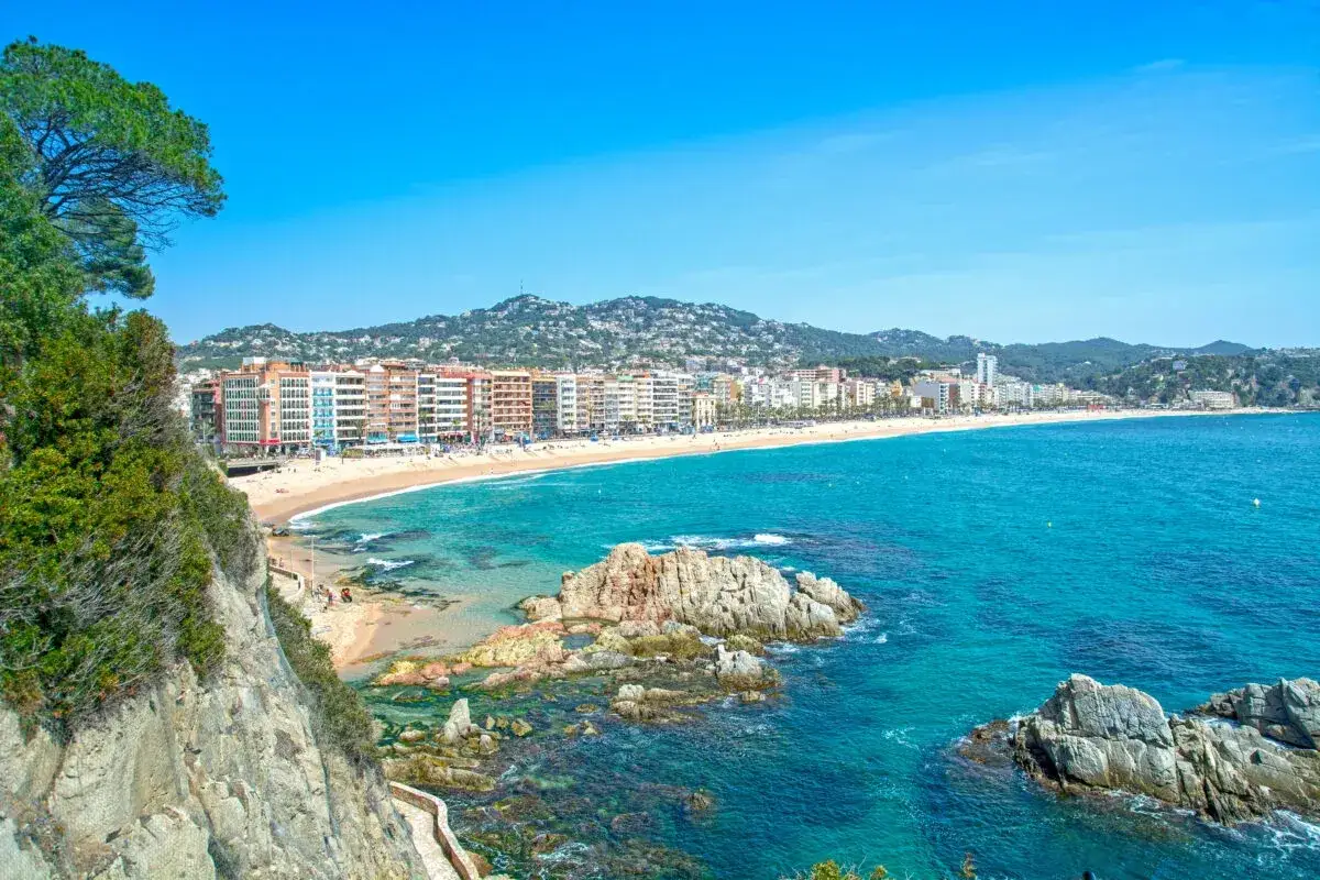 Widok na plażę w Lloret de Mar, z turkusowym morzem i skalistymi zatoczkami. W oddali widać zabudowania miasta, a wzgórza pokryte zielenią. Idealne miejsce na wakacje, niedaleko od lotniska.