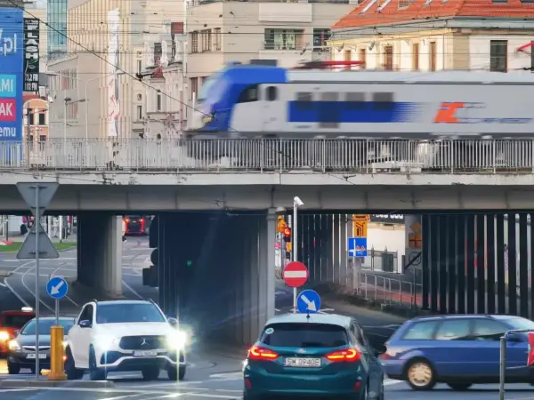 Pędzący pociąg IC 407 na trasie Chopin mija ruchliwe skrzyżowanie z samochodami i tramwajami w tle.